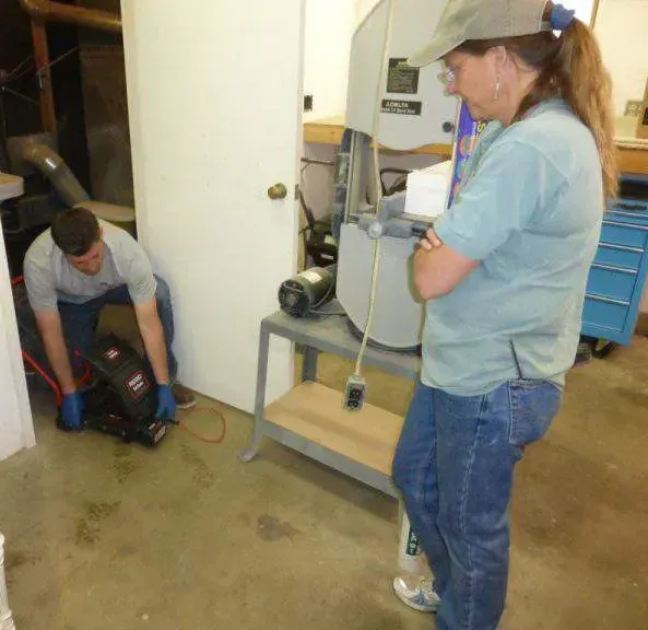 A person kneels on a concrete floor to inspect a piece of equipment while another person stands and observes.