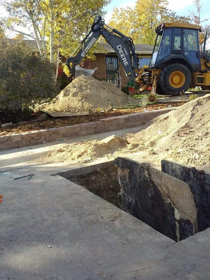 A John Deere backhoe sits in a residential yard near a pile of excavated dirt and an open trench in a concrete driveway.