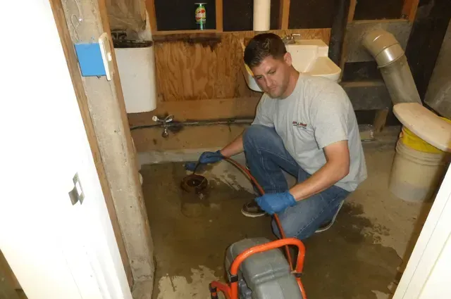 A person in a gray shirt and blue gloves uses a plumbing auger to clear a floor drain in a utility room.