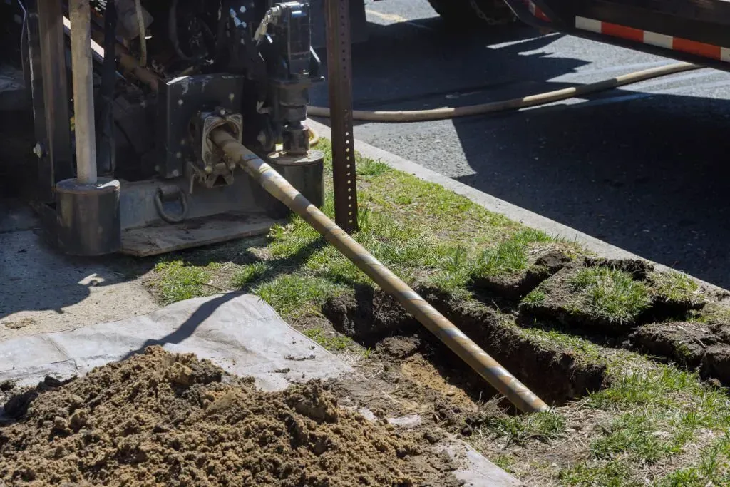 A machine drills a metal rod into the ground at an angle near a roadside patch of grass and a pile of dirt.