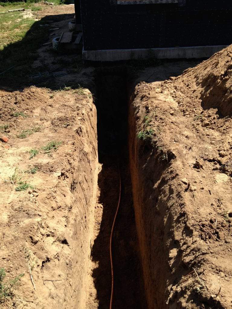A deep, narrow trench dug into dirt alongside the foundation of a building, with a utility cable running along the bottom.
