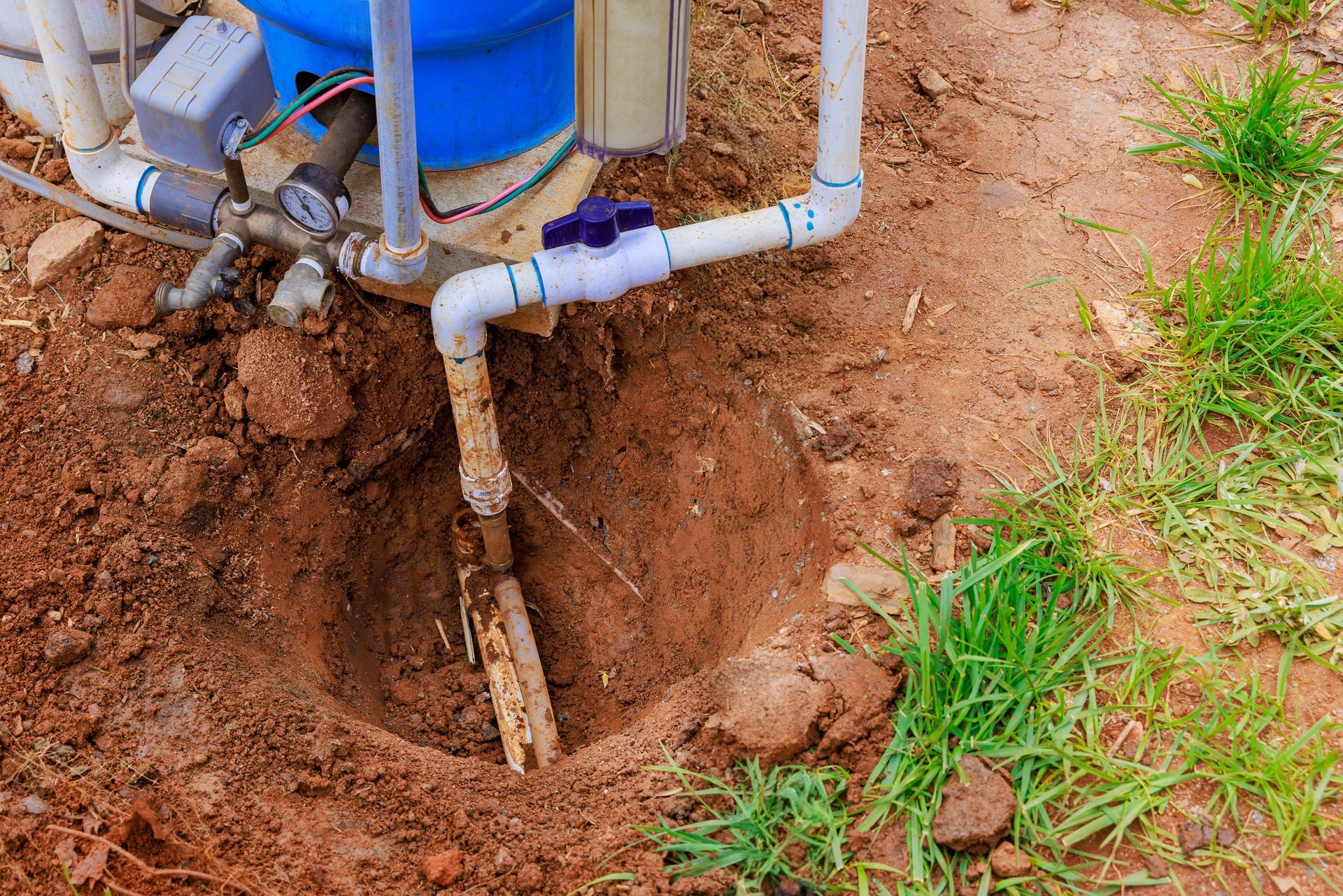 Hole dug around water pipes, near a blue tank and white PVC pipes. Brown soil, some grass.