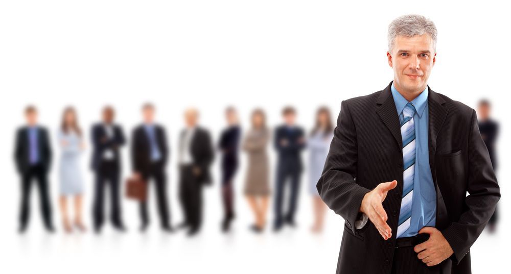 Man in suit extends hand to greet, other professionals blurred in the background.