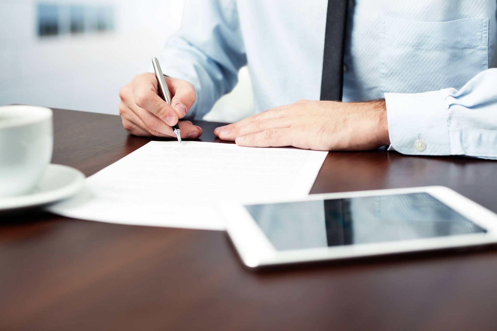 Person in blue shirt signing a document with a pen at a wooden desk with a tablet and coffee cup.