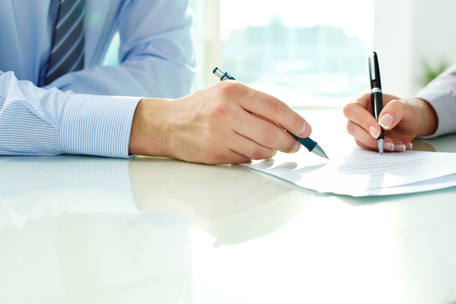 Two people signing documents at a white table, one wearing a blue shirt and tie.