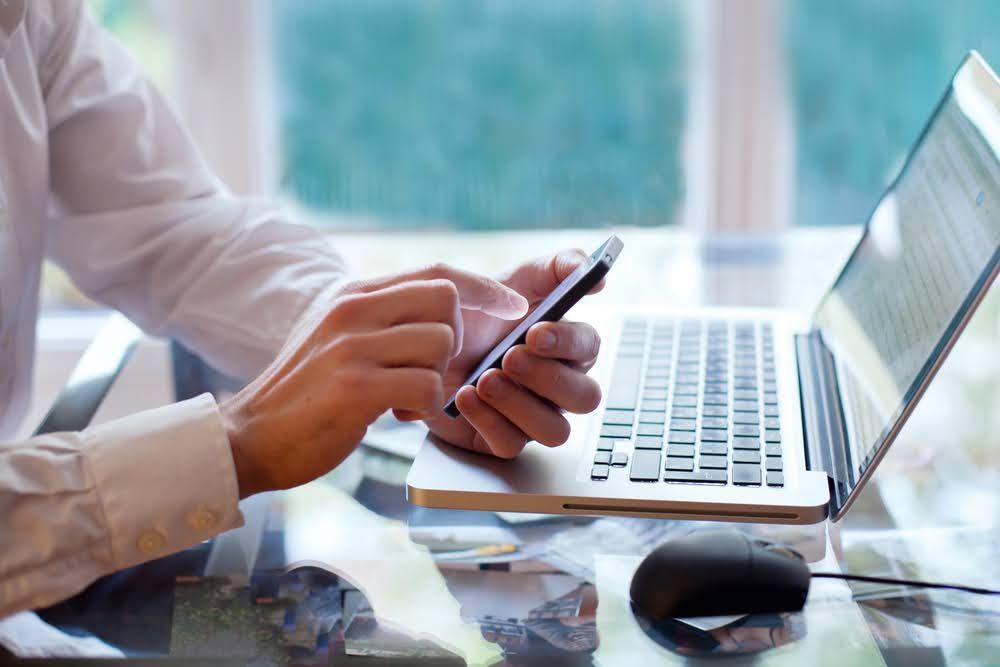 Person using a smartphone and laptop at a desk with a mouse.