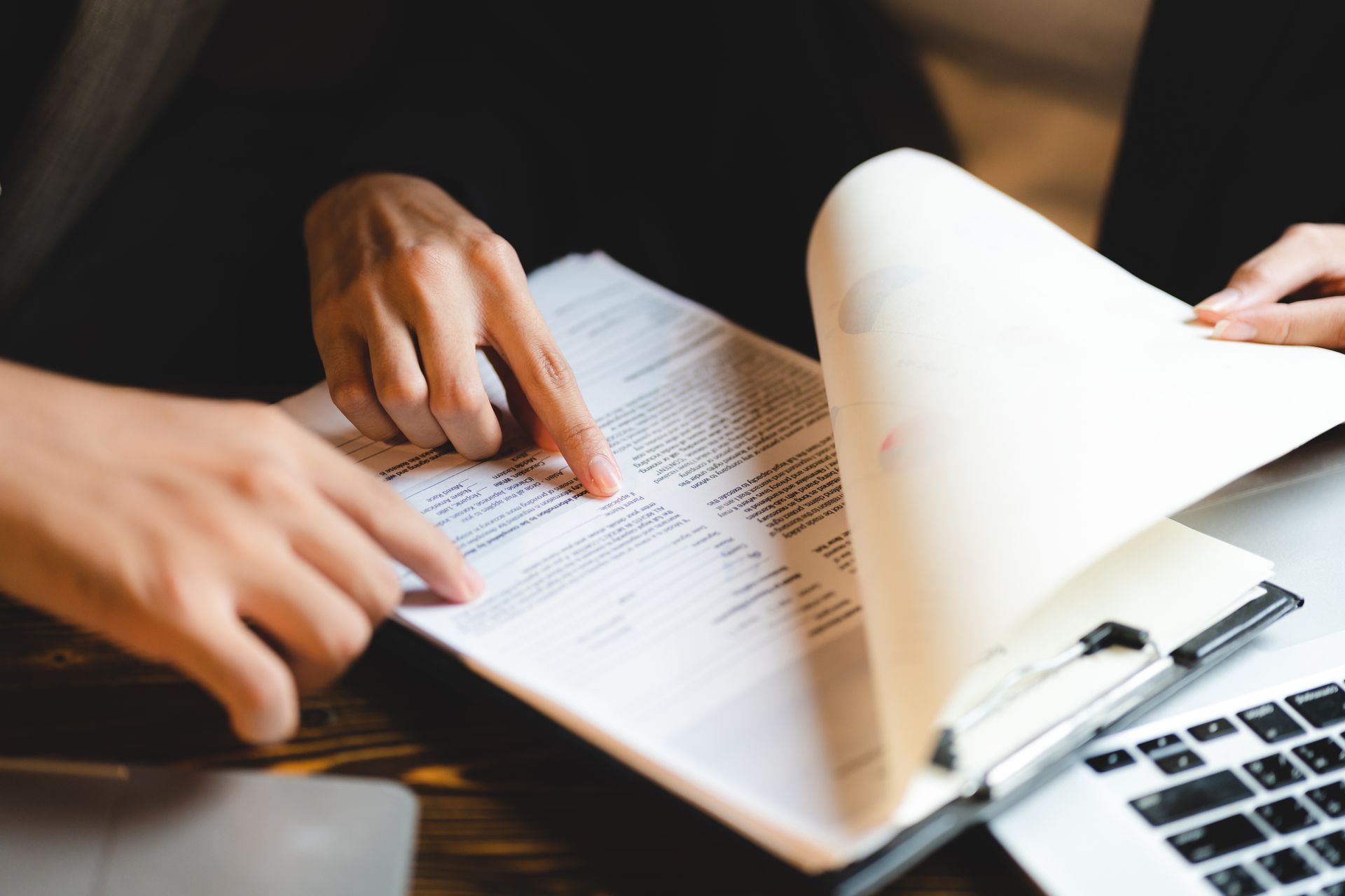 Hands reviewing a document beside a laptop, with one page being turned over Hands reviewing a document beside a laptop, with one page being turned over