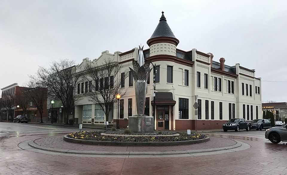 Corner building with a statue in a roundabout on a wet street. Cars parked on the side. Overcast sky.