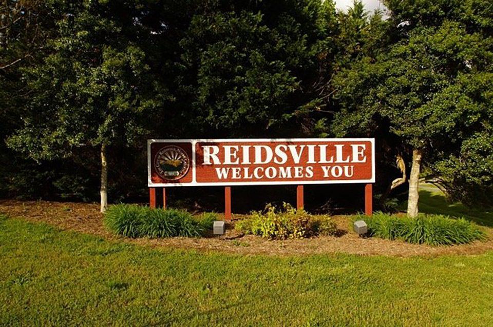 Sign welcoming visitors to Reidsville, brown with white lettering, in front of trees and grass.