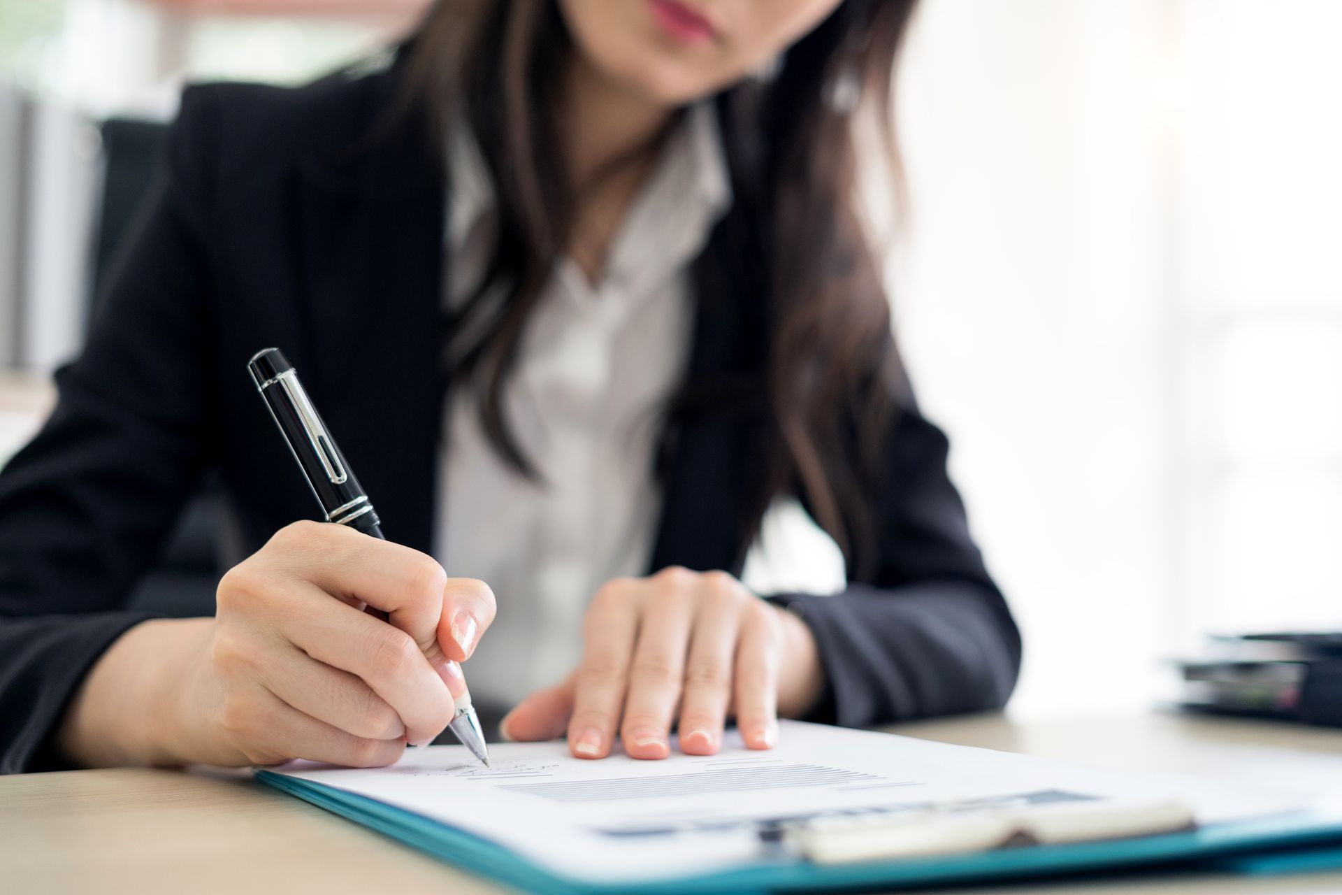 Woman in black blazer signing document with pen at a desk.