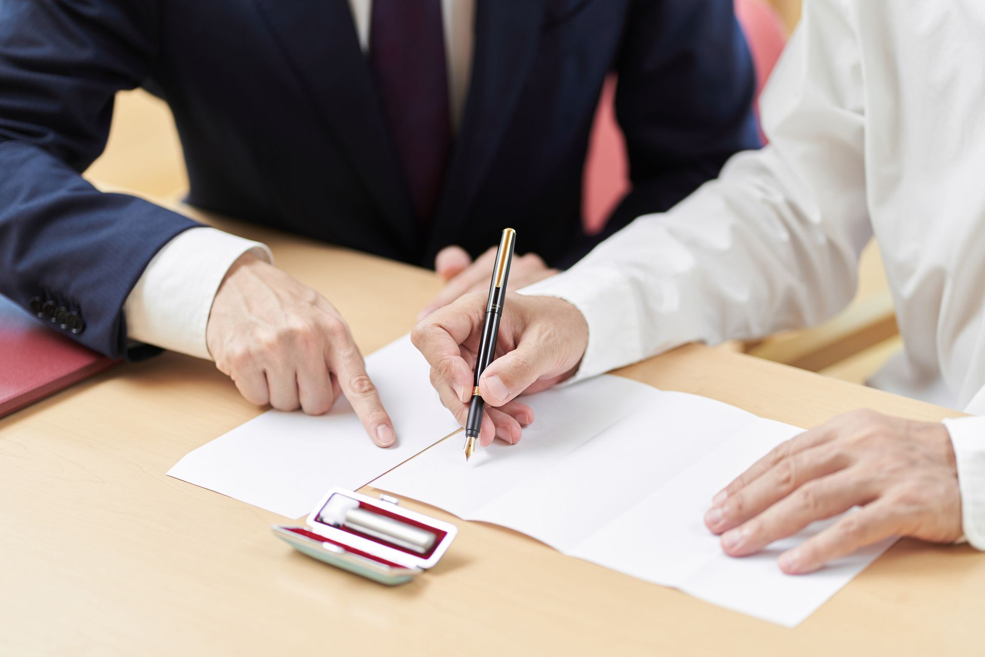 Two men at a desk, one points to a document while the other signs it with a pen.
