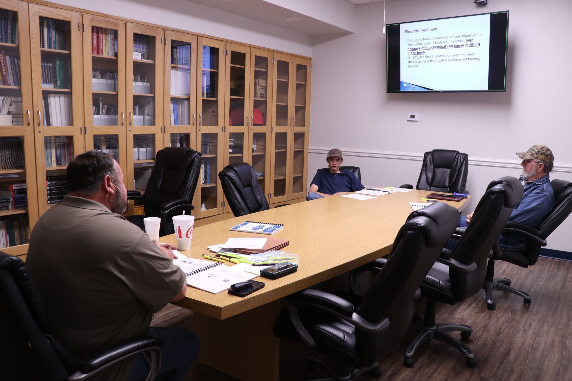 Small conference room with people seated around a table, facing a wall-mounted presentation screen.