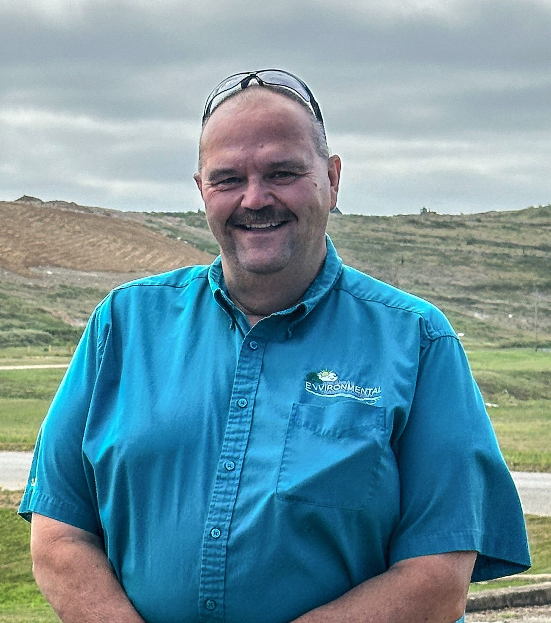 Man smiling outdoors in a blue shirt, standing in front of a grassy landscape and cloudy sky.