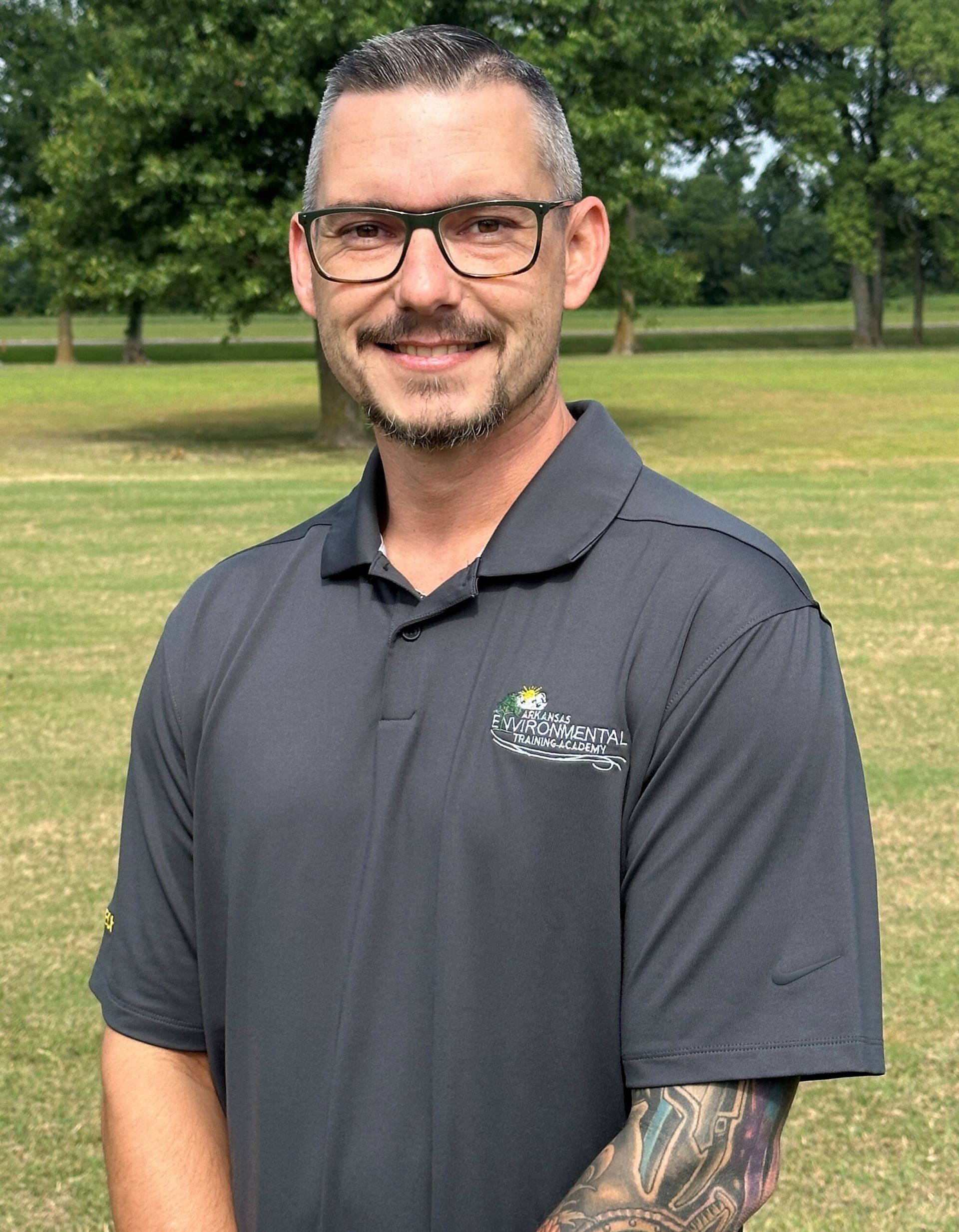 Smiling man in black polo and glasses standing outdoors on a grassy field.