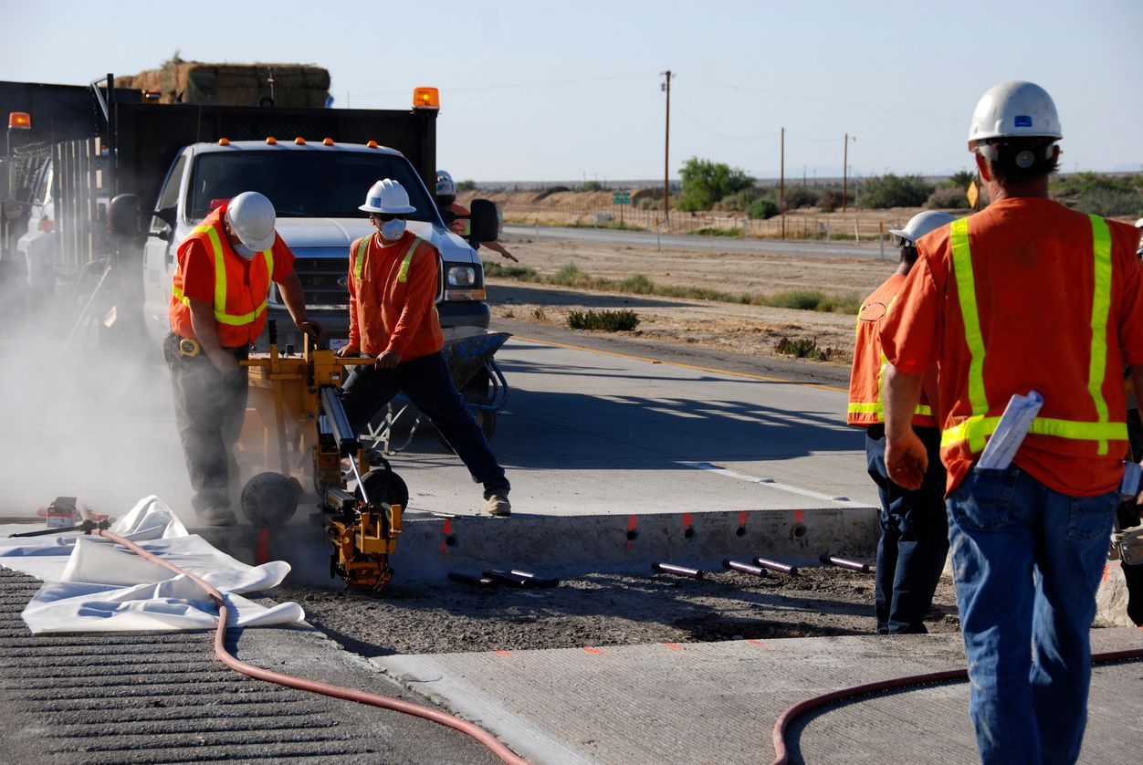 Construction workers in orange safety vests repair a road beside a truck, with steam or dust rising.