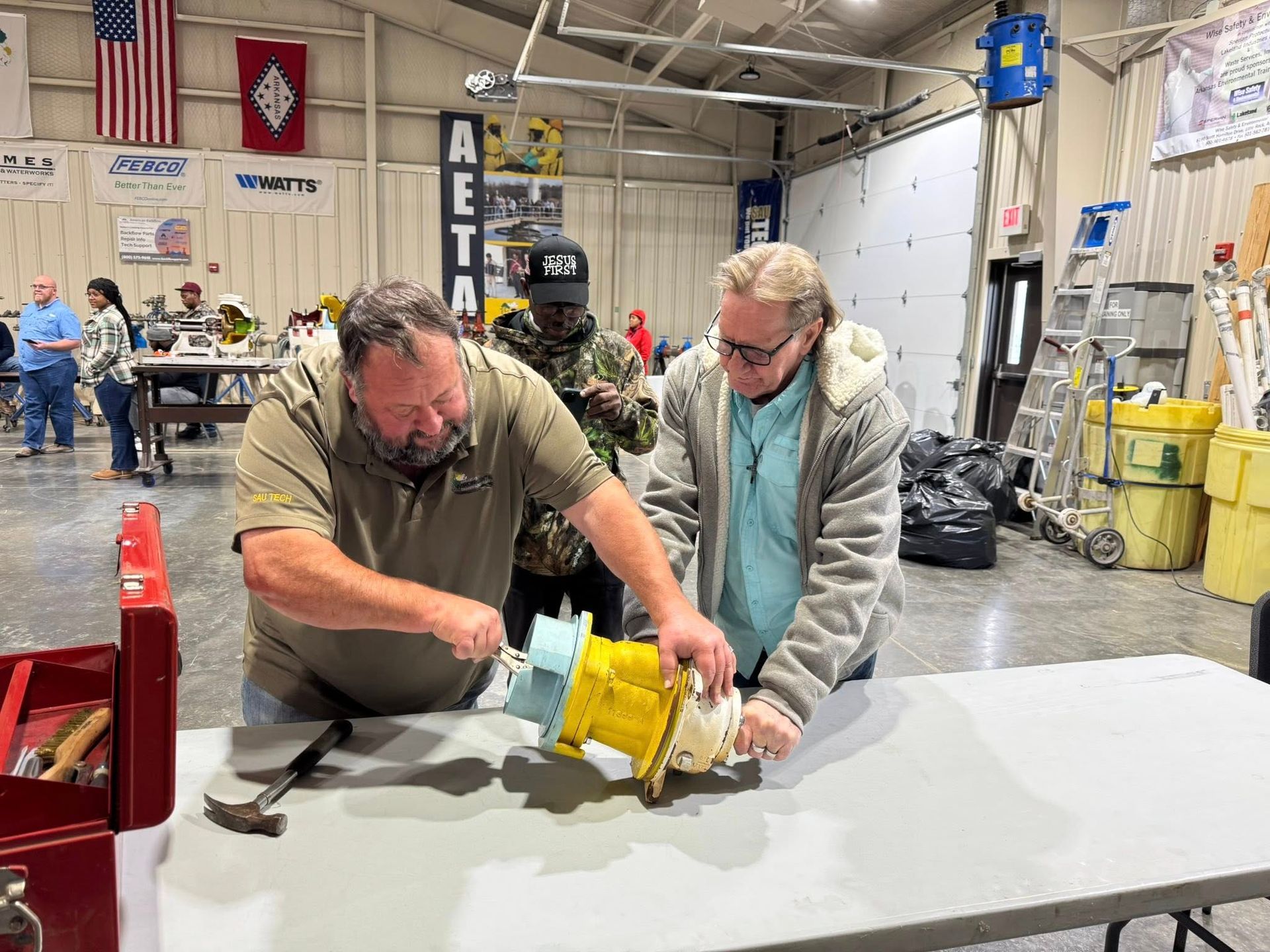 Two people working with tools at a workbench in a workshop, handling a yellow machine part