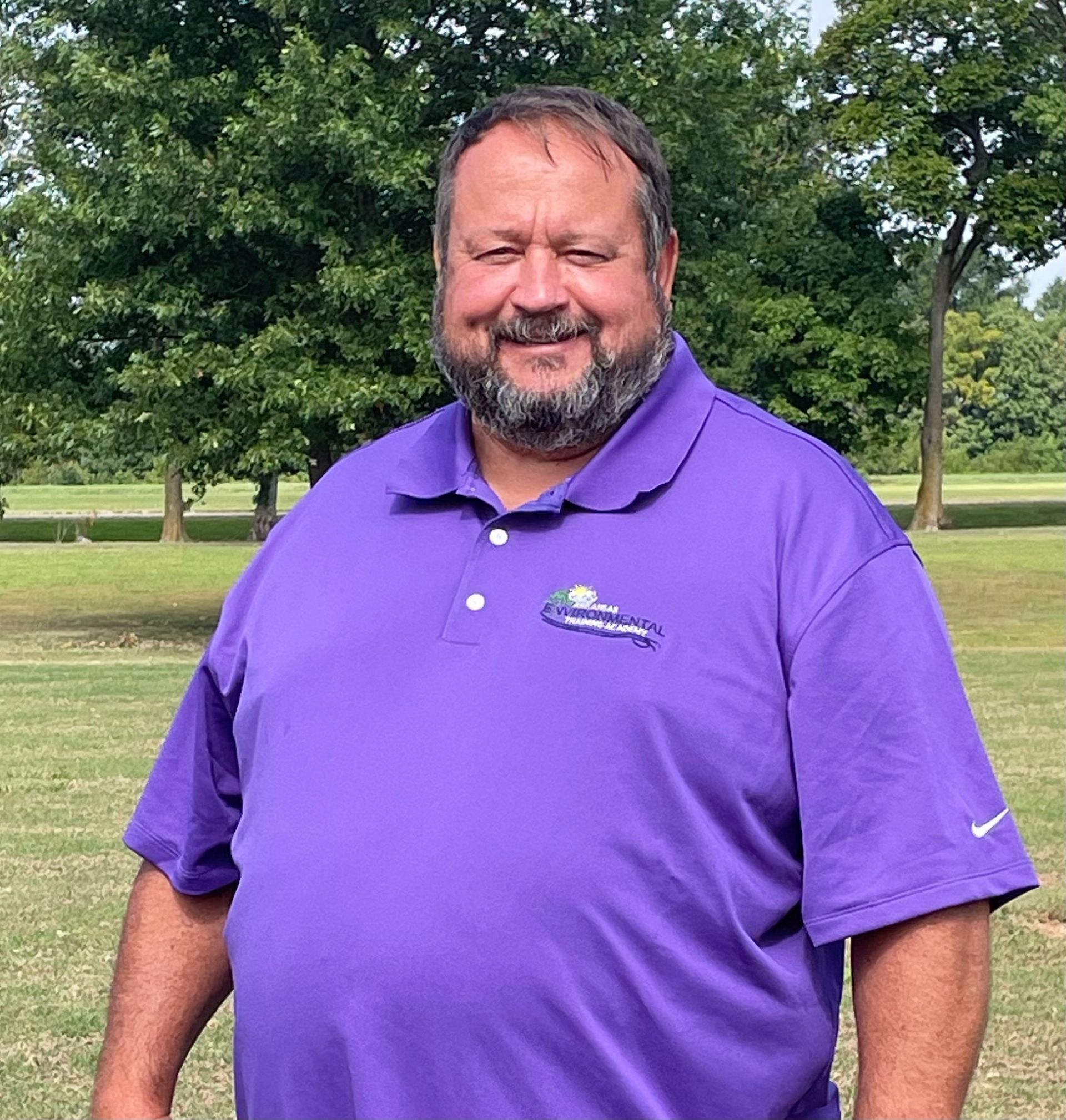 Man in a purple polo shirt smiling in a park with trees and grass in the background