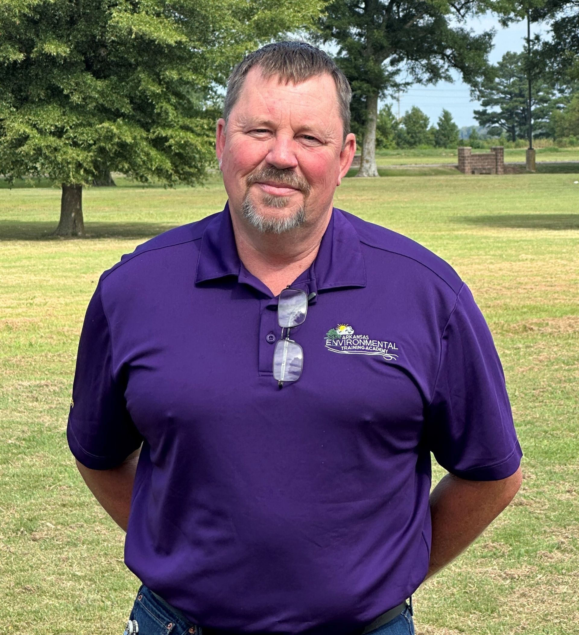 Man in a purple polo standing outdoors in a grassy park, hands behind his back.
