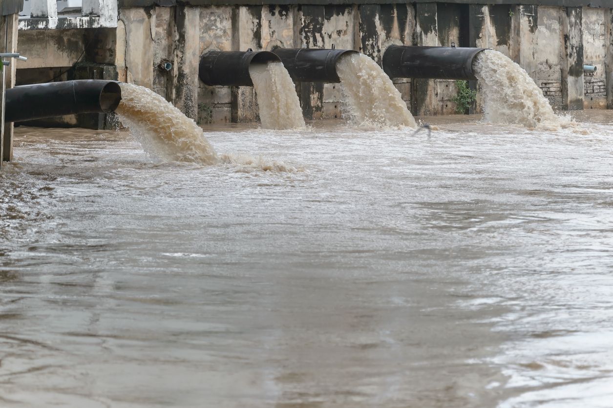 Wastewater pipes pouring water into a choppy, muddy channel beside a concrete wall