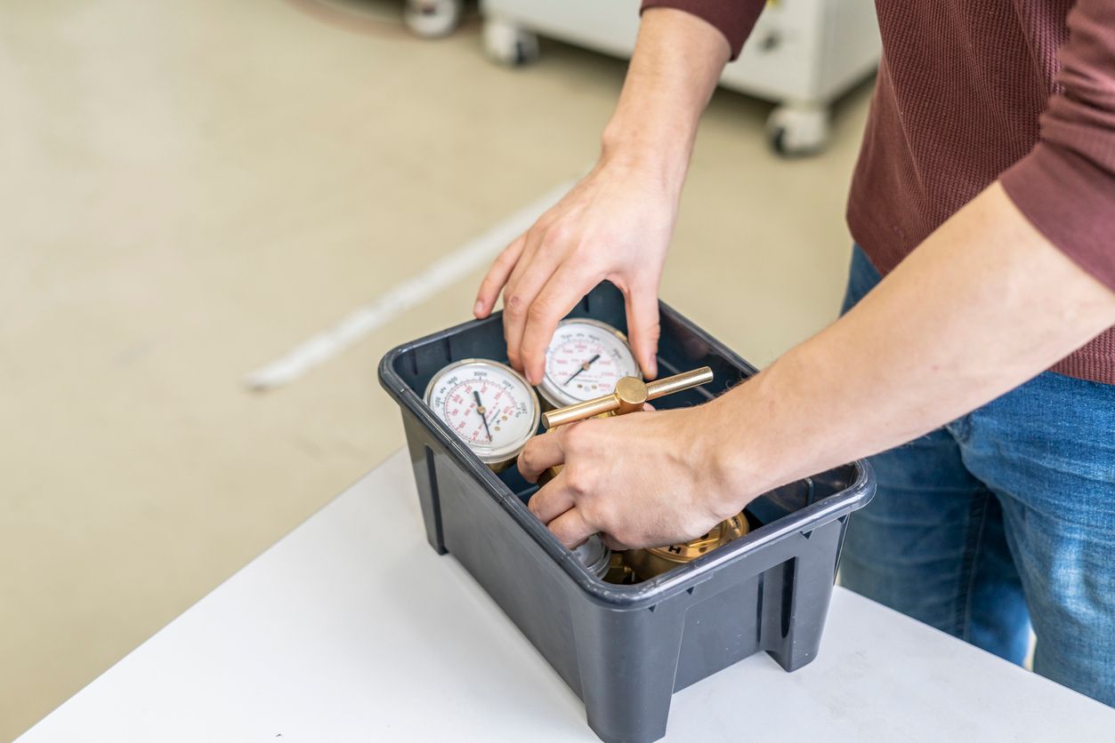 Hands placing items into a black crate on a table beside two pressure gauges.