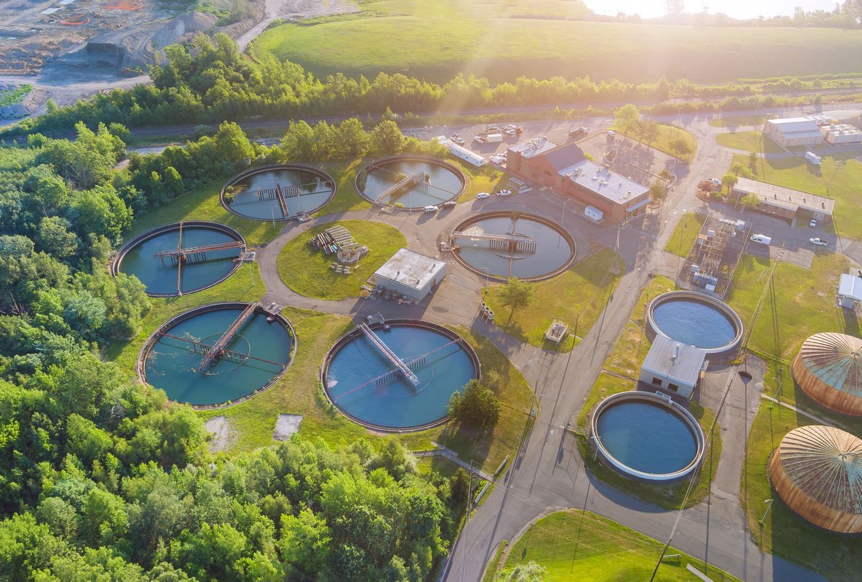 Aerial view of a circular wastewater treatment facility with multiple blue basins surrounded by green trees.