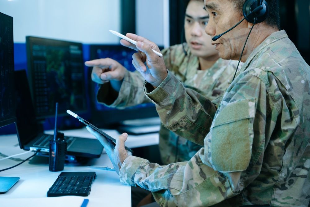 Two military personnel at computers in a control room, one pointing at a monitor while wearing a headset.