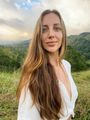 Woman with long brown hair in a white dress, smiling in a lush green hillside landscape at sunset