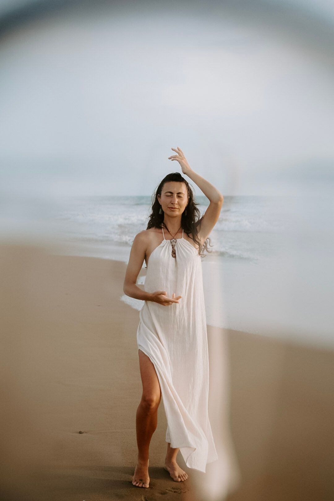 Woman in a flowing white dress standing on a beach with waves behind her, one arm raised overhead.