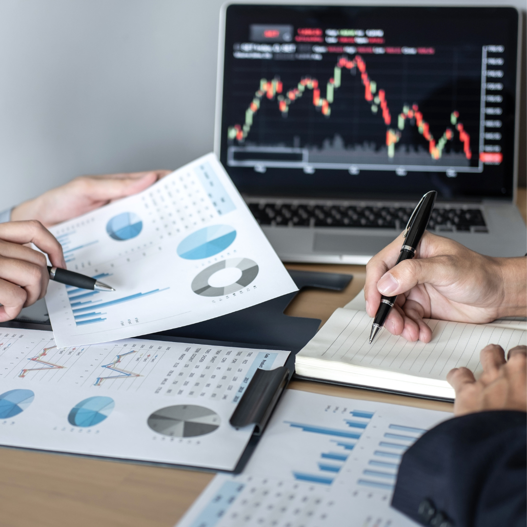 Hands reviewing financial data on paper, with a laptop showing a stock chart.