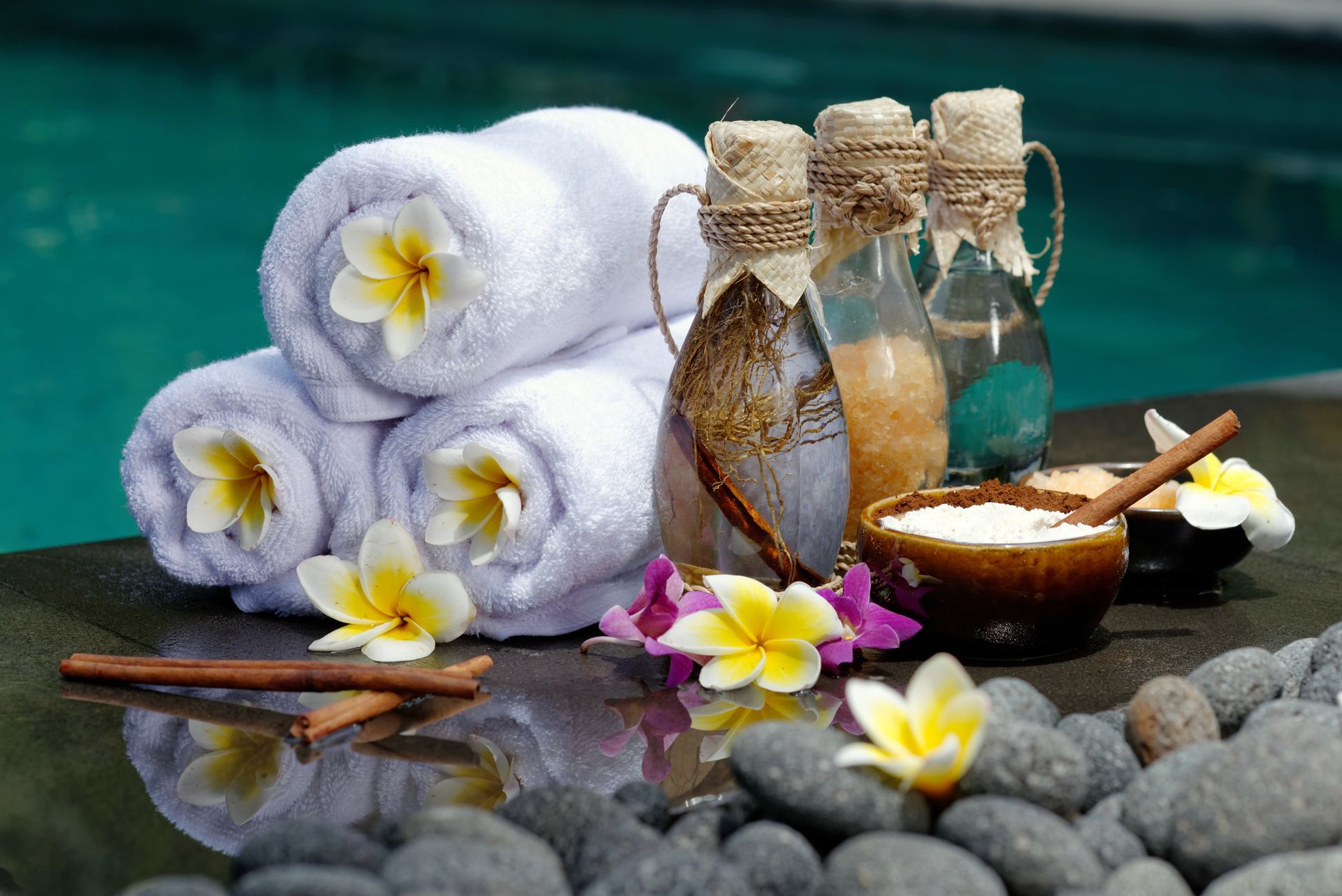 Allestimento spa con asciugamani bianchi arrotolati, bottiglie, ciotole e fiori a bordo piscina.