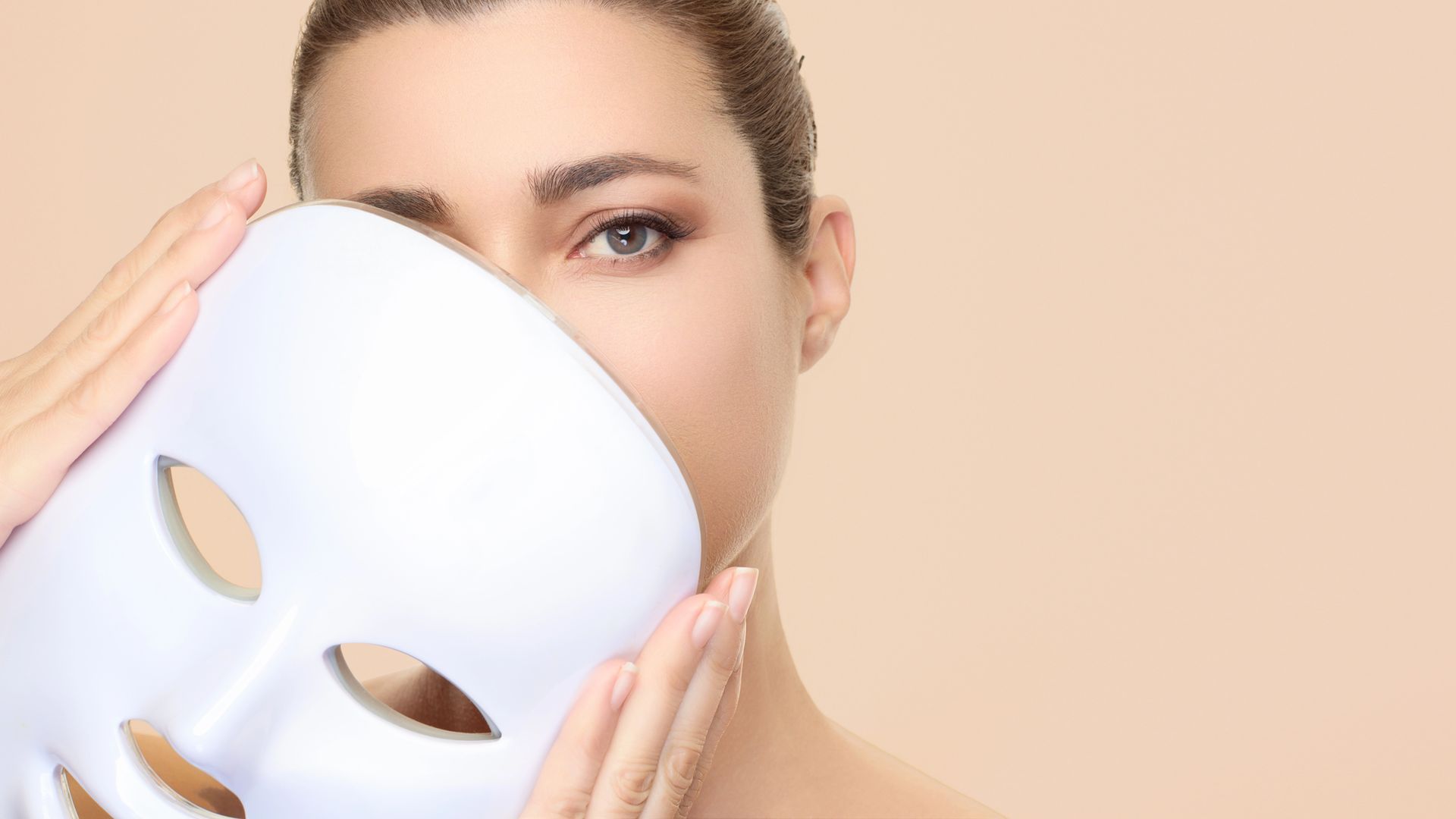 Donna che tiene una maschera per la terapia con luce LED bianca parzialmente coperta sul viso. Sfondo color crema.
