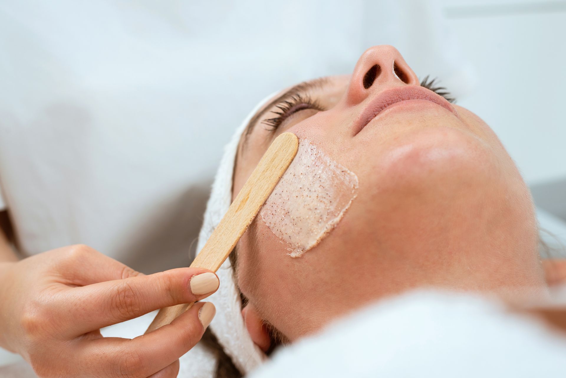 Persona che riceve un trattamento viso; estetista che applica il prodotto con una spatola di legno.