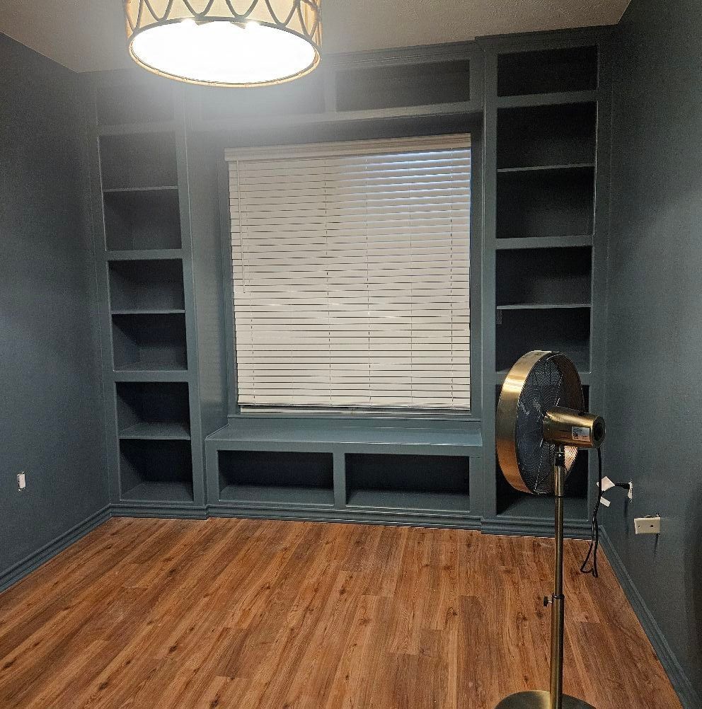 Built-in shelves frame a window with a bench below. A fan stands near the window, and the walls are dark blue.
