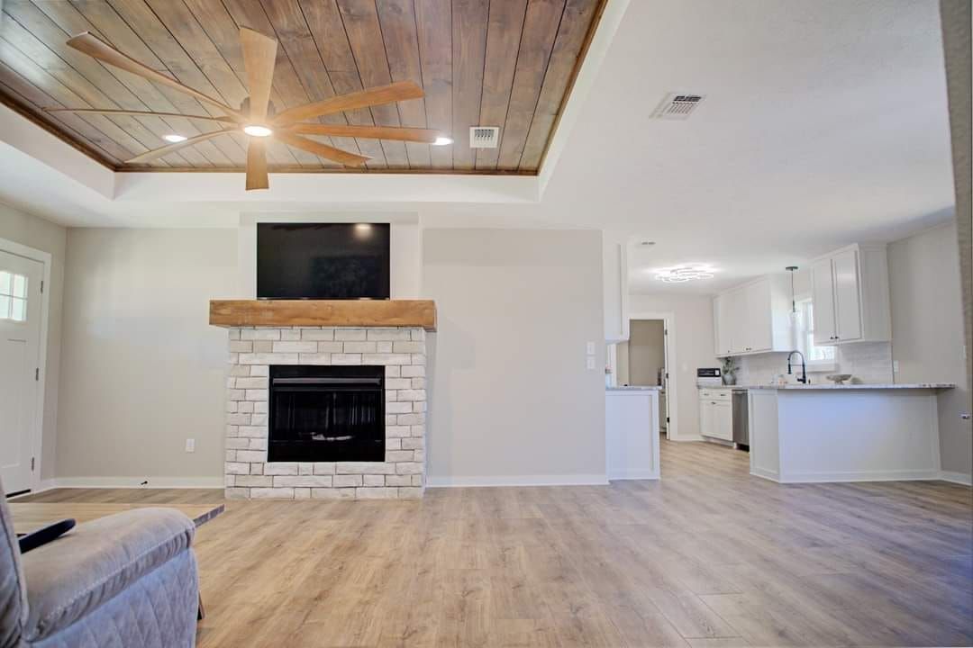 Living room with fireplace, TV, wood ceiling, light-colored walls and floor, and partially visible kitchen.