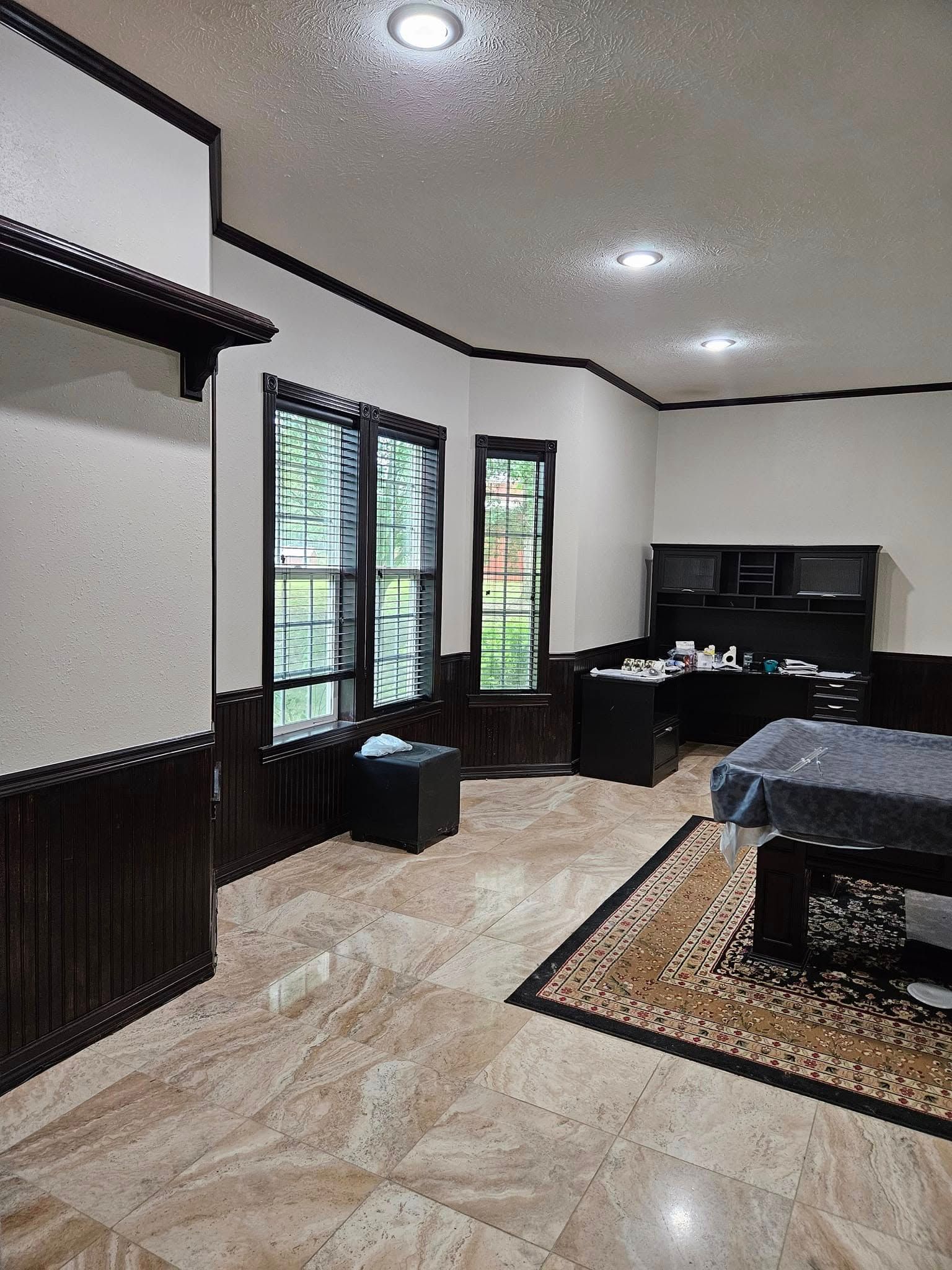 Office interior with dark wood trim, light walls, and tiled floor; a desk, windows, and rug are visible.