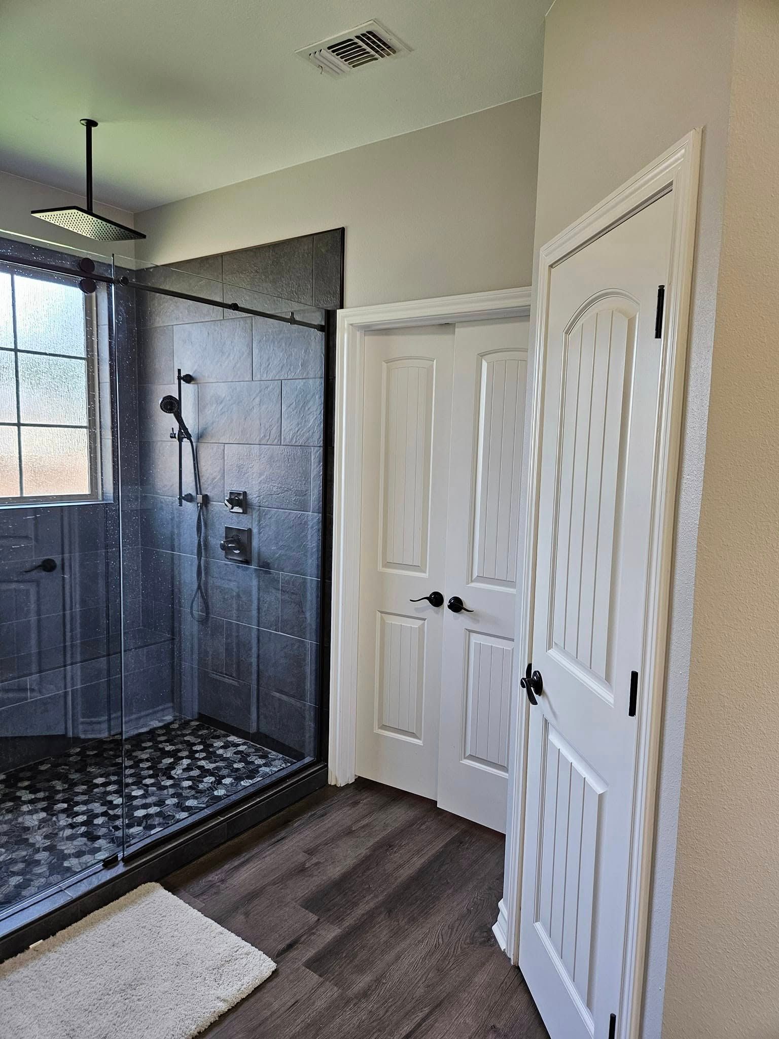 Bathroom with dark gray shower, pebbled floor, two white doors, and dark wood-look floor.