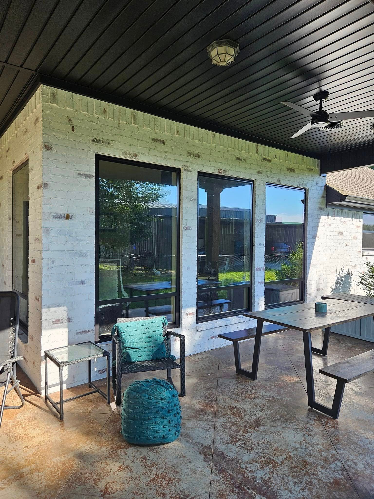 Outdoor patio with brick walls, large windows, and a black ceiling. Furniture includes a chair, ottoman, and table.