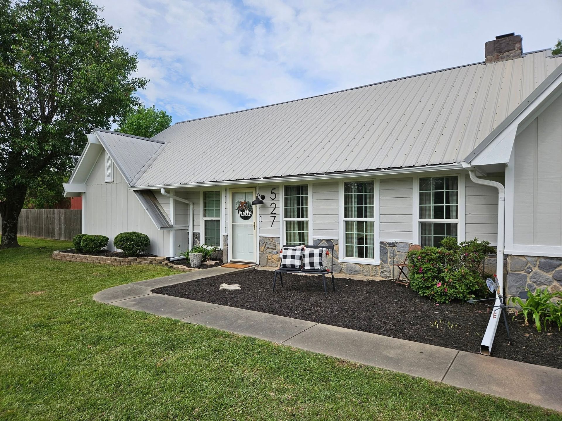 White house with metal roof, stone accents, and manicured landscaping, under a cloudy sky.