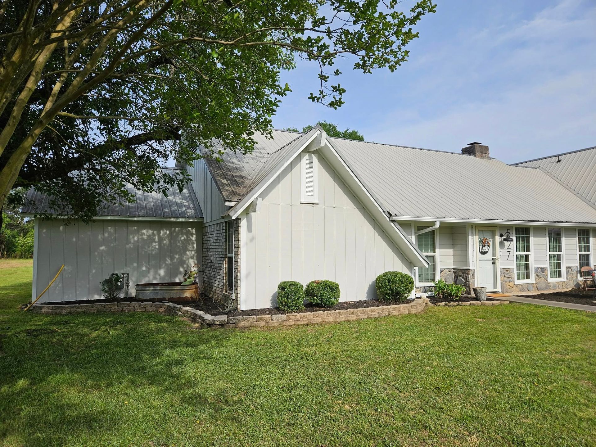 White house with gray roof, green lawn, tree on the left, bushes and landscaping in front.