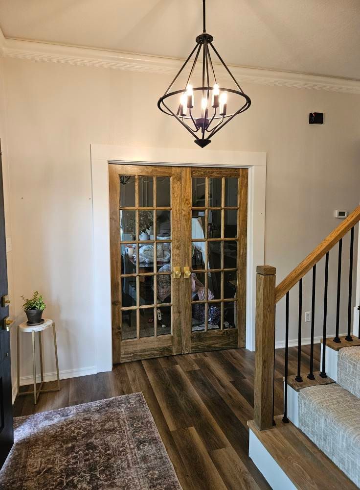 Entryway with wood-paneled double doors, chandelier, and staircase. Features wood flooring and a small table with plant.