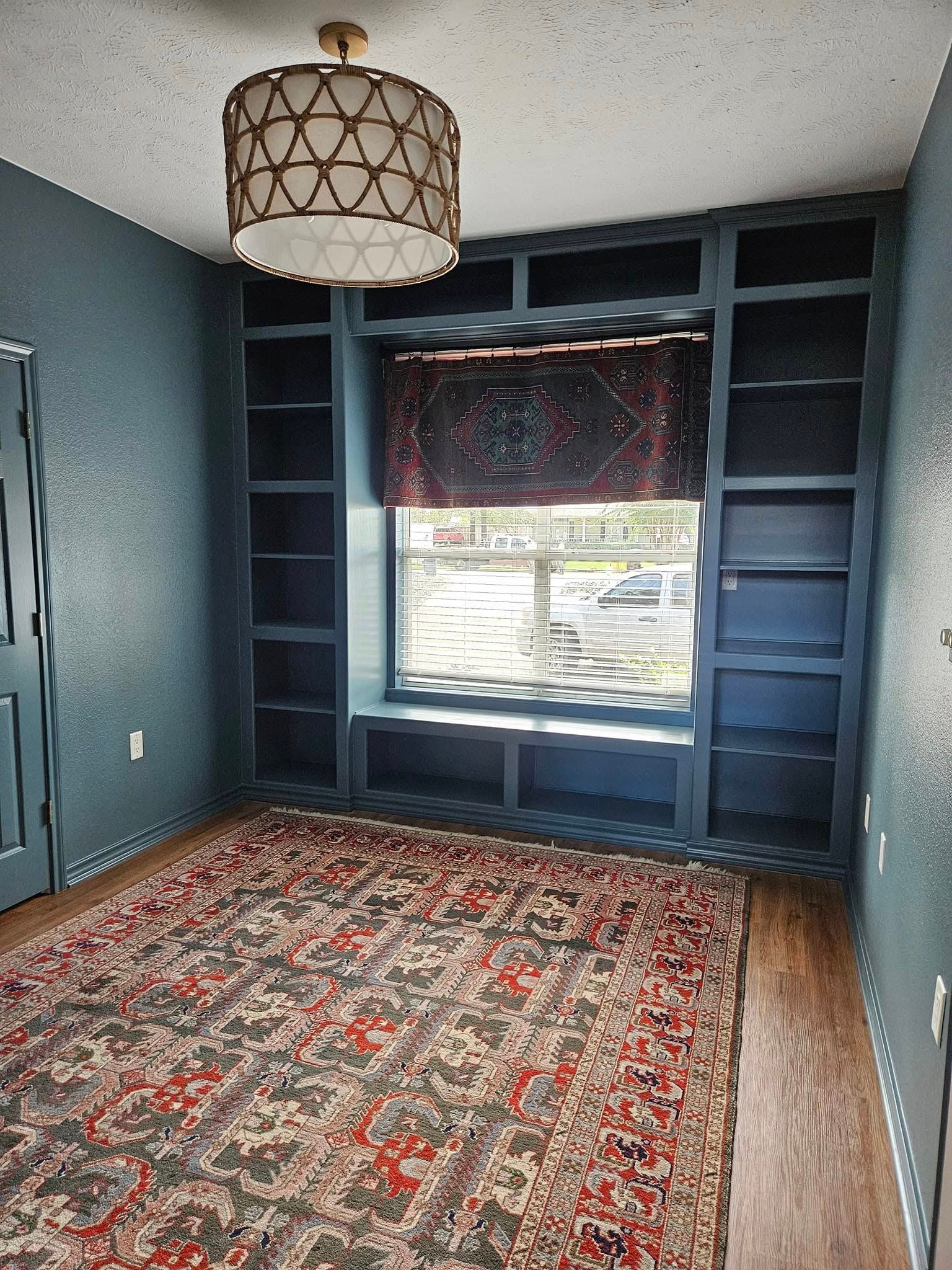 Room with built-in bookshelves painted teal, window seat with patterned shade, oriental rug, wood floor, and hanging lamp.