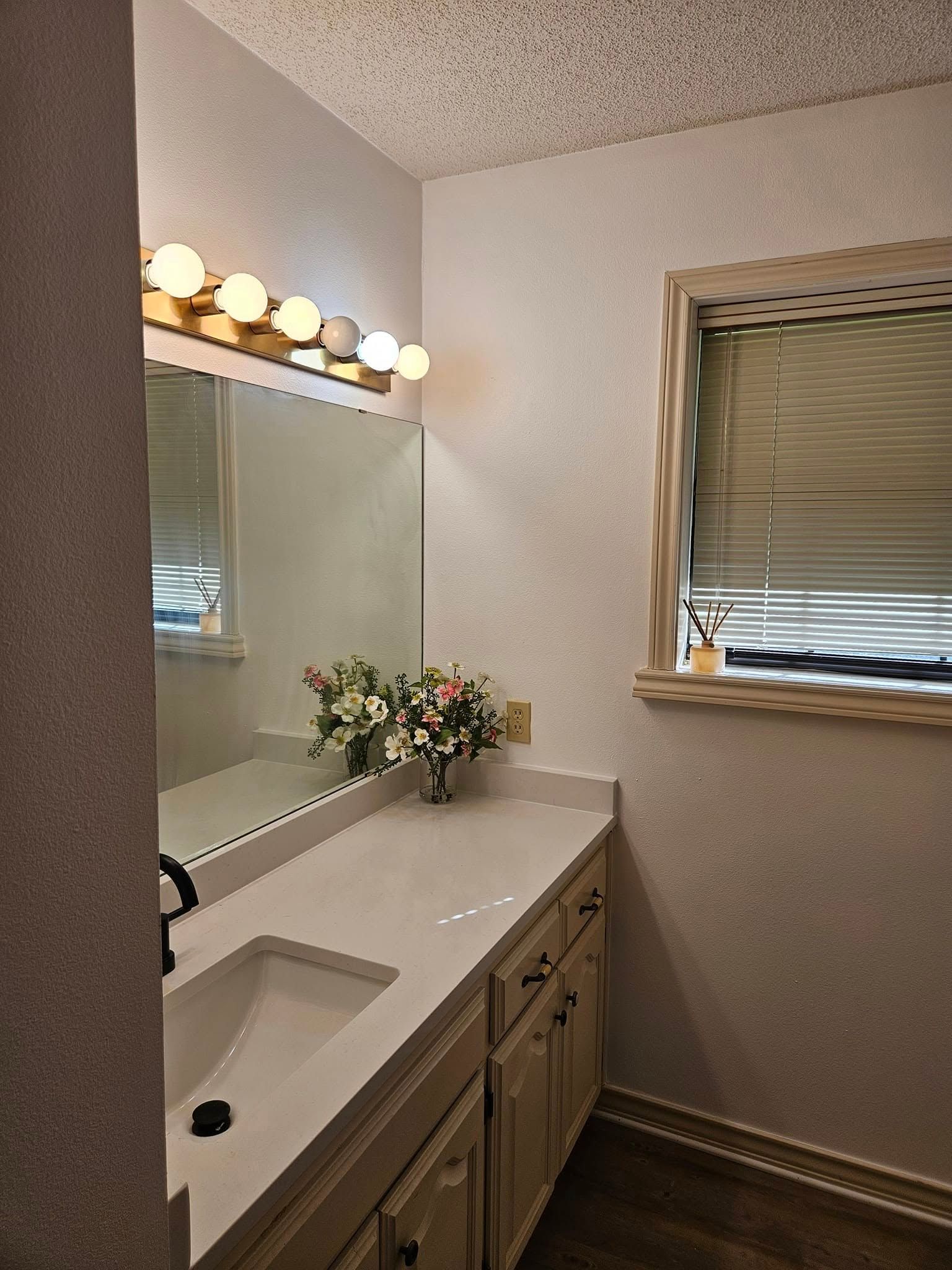 Bathroom with white countertop, mirror, vanity, and window with blinds. A vase of flowers sits on the counter.