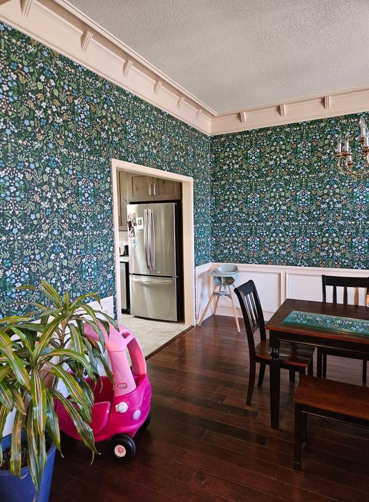 Dining room with patterned teal wallpaper, dark wood floors, and a glimpse into the kitchen.