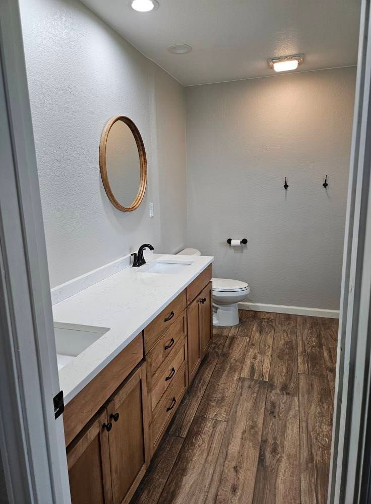 Bathroom with wood cabinets, white countertop, and wood-look floor. A round mirror hangs above the sink.