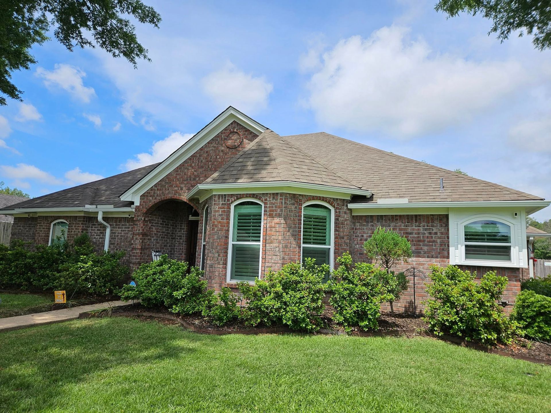 Brick house with a dark roof and green bushes under windows. Blue sky overhead.