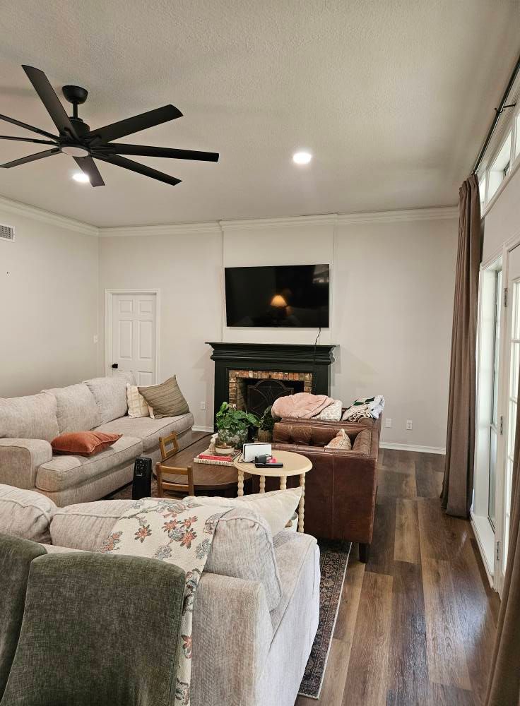 Living room with couches, fireplace, TV, and ceiling fan. Dark wood floors and neutral walls.