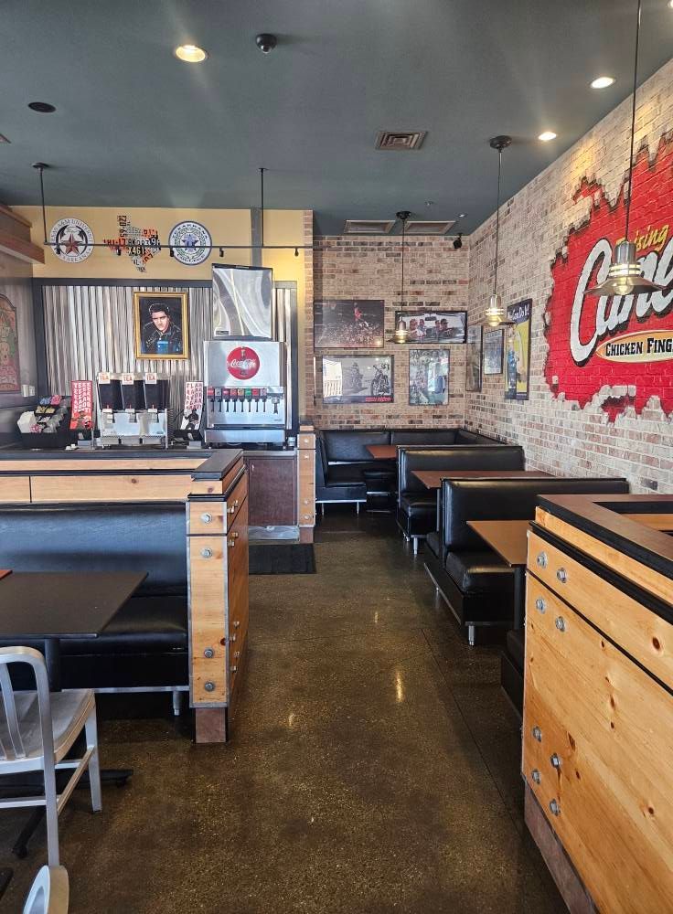 Interior of a restaurant with booths and tables. Red brick wall with logo, concrete floor, counter with beverage dispensers.