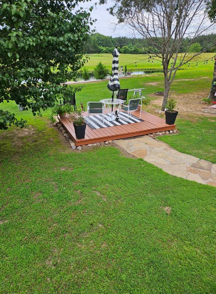 Outdoor deck with table, chairs, striped rug, umbrella, and potted plants on a grassy lawn.