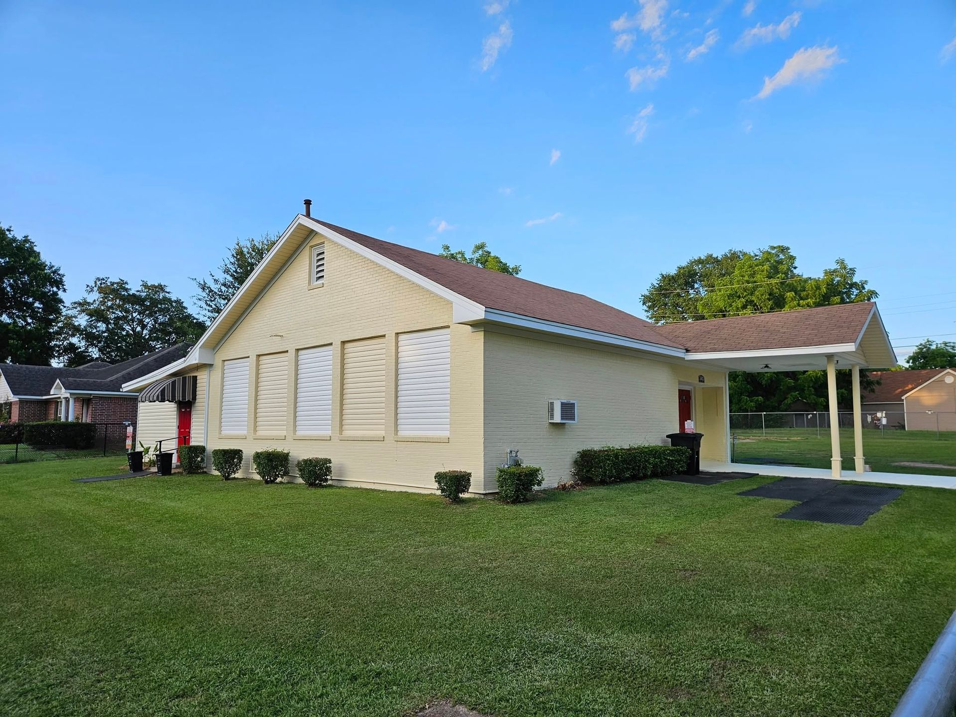 Tan one-story building with brown roof, carport, and green lawn under a blue sky.