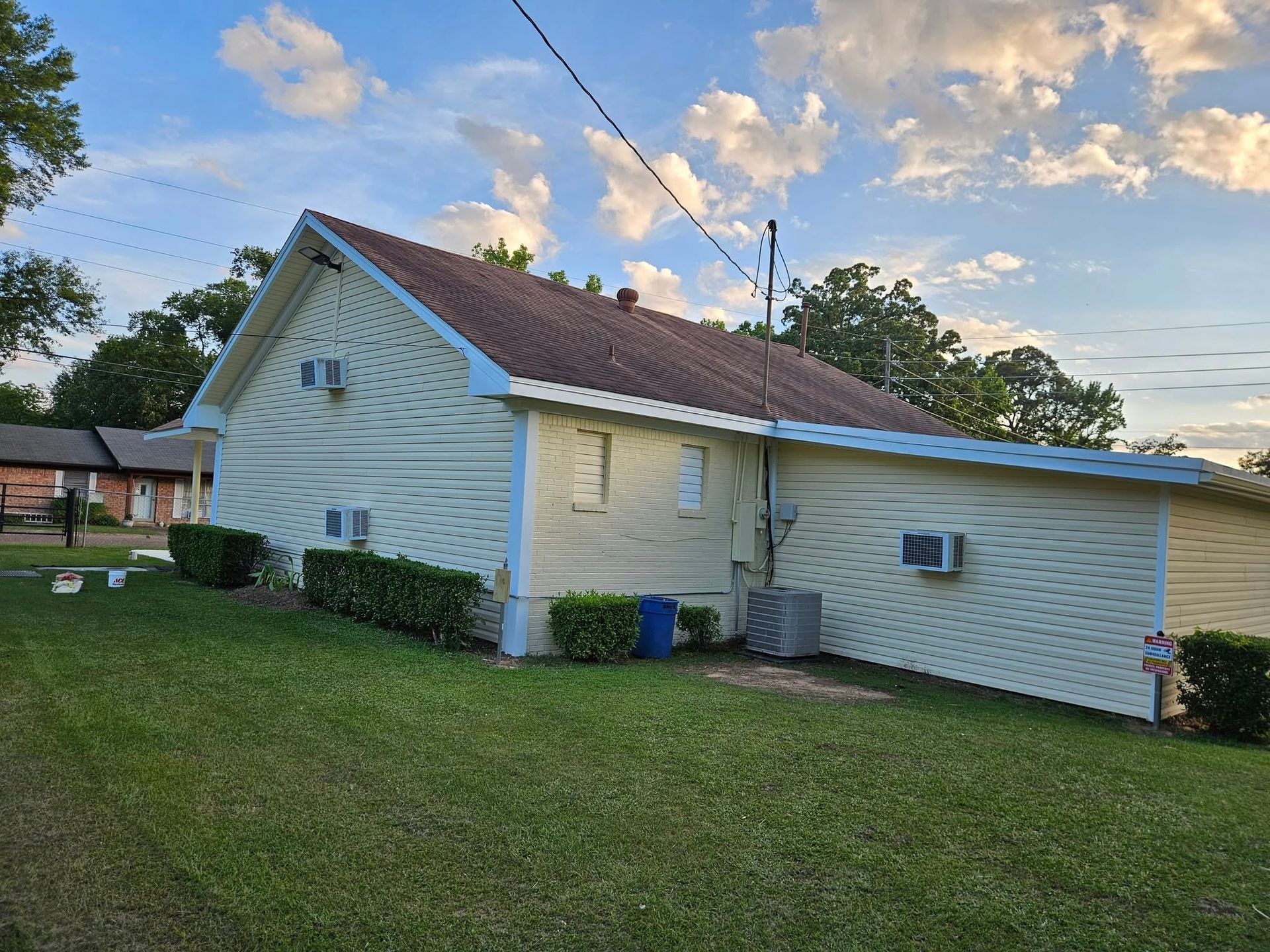 Yellow house with brown roof, air conditioning units, and green bushes against a cloudy sky.